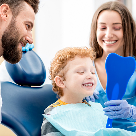 Little boy checking smile in mirror with dentist and parent nearby