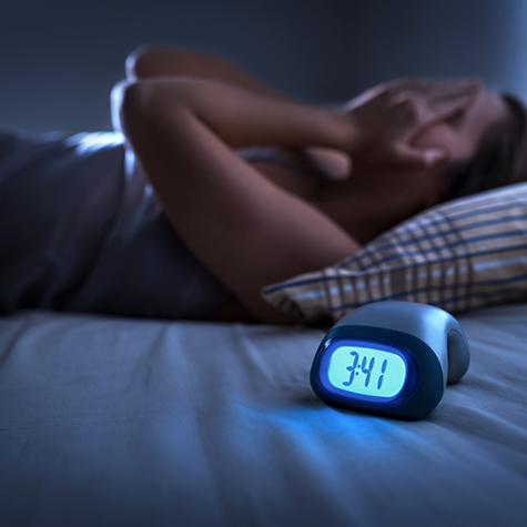 Woman lying on bed with hands over face with clock in foreground